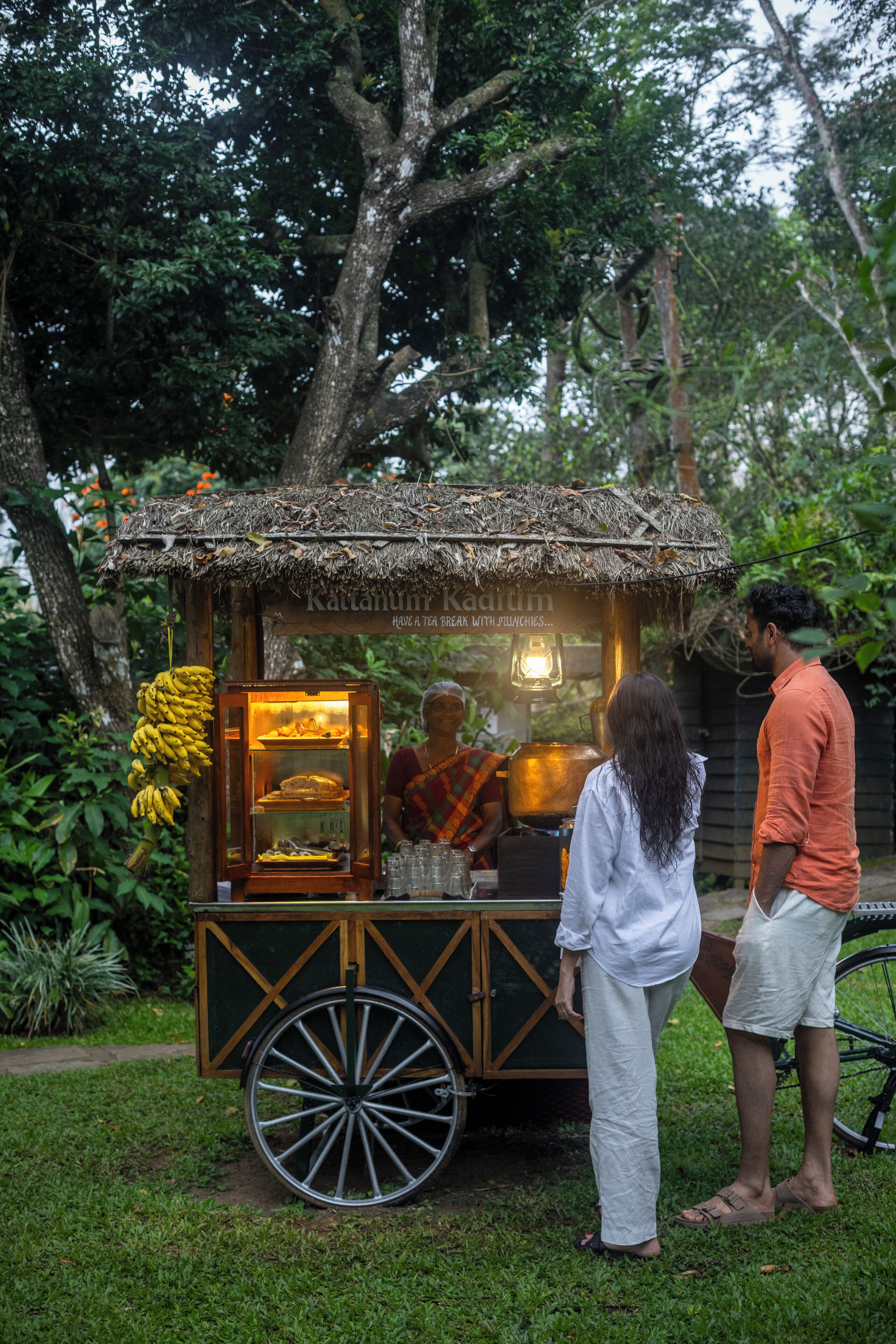 A food cart with a thatched roof, bananas hanging on the side, and a person serving food.