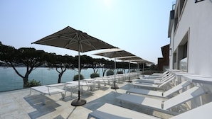 Una terraza con sillones blancos y grandes sombrillas, con vista a un cuerpo de agua y árboles.