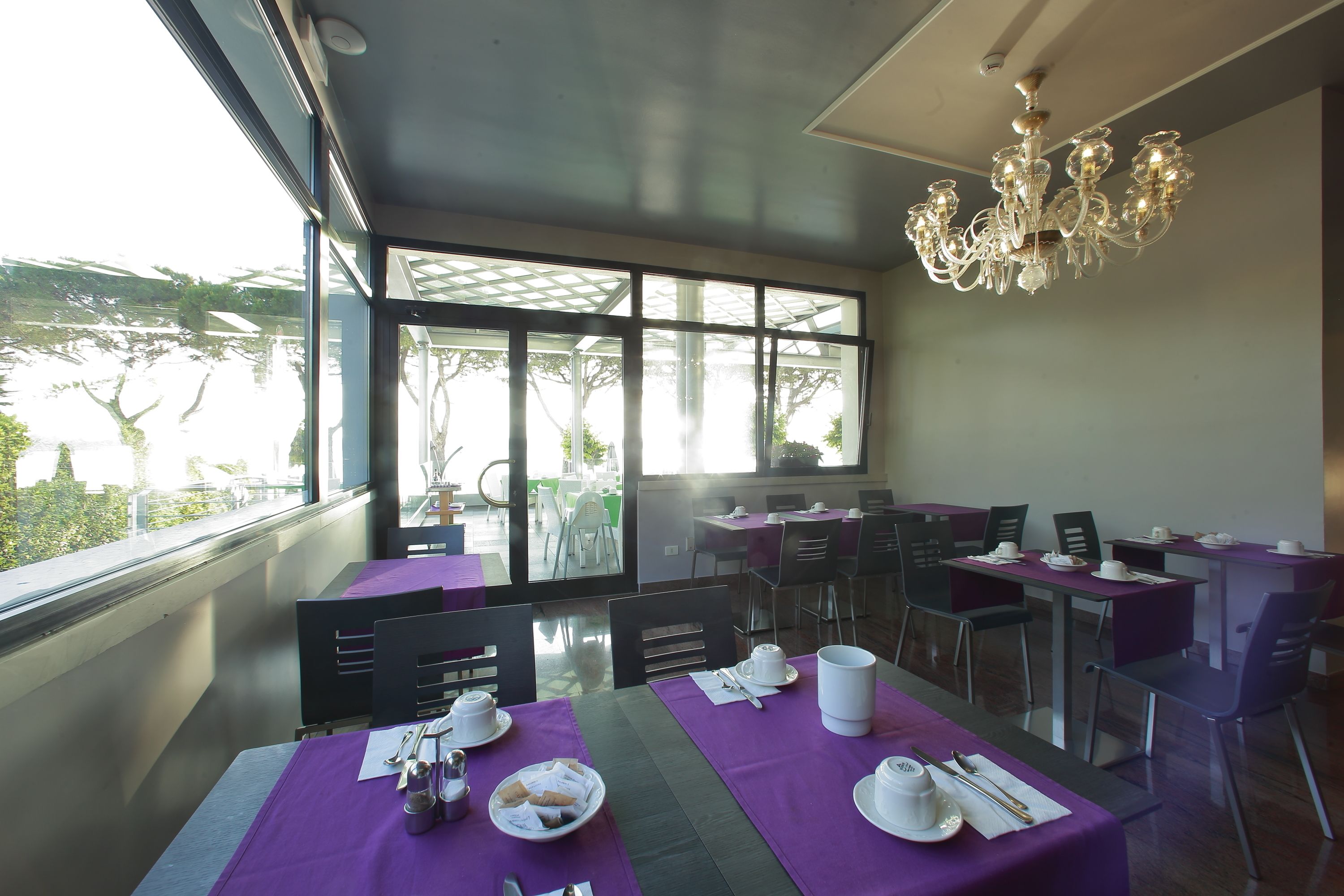 A dining area with tables set for breakfast, large windows, and a chandelier.