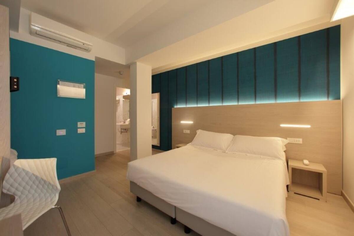 A modern hotel room with a large bed, a bedside table, a chair, and a bathroom visible through an open door.