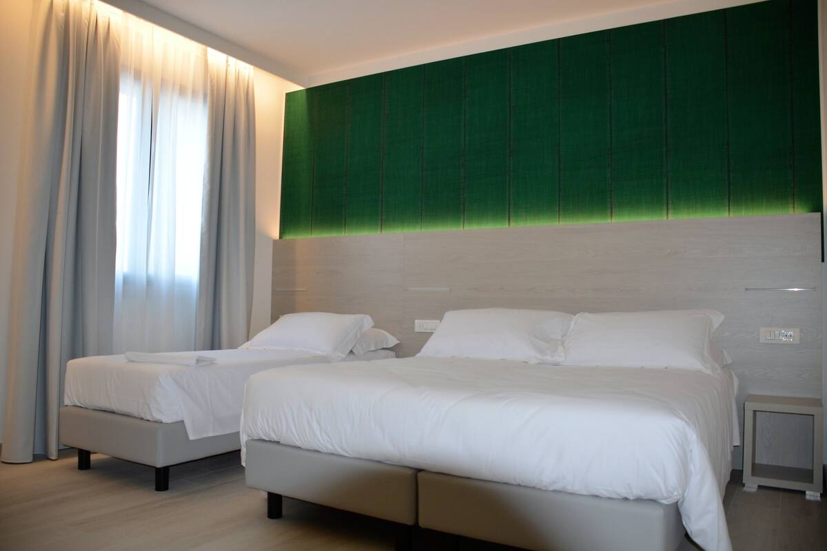 A modern hotel room with two beds, a green accent wall, and a large window with curtains.