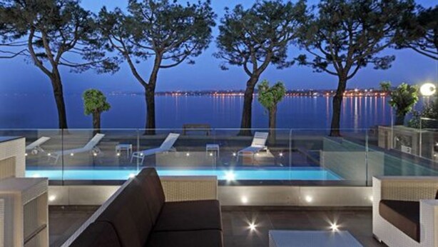 Una piscina infinita con sillones de descanso y barandilla de vidrio, con vistas a una masa de agua y el horizonte de una ciudad al anochecer.