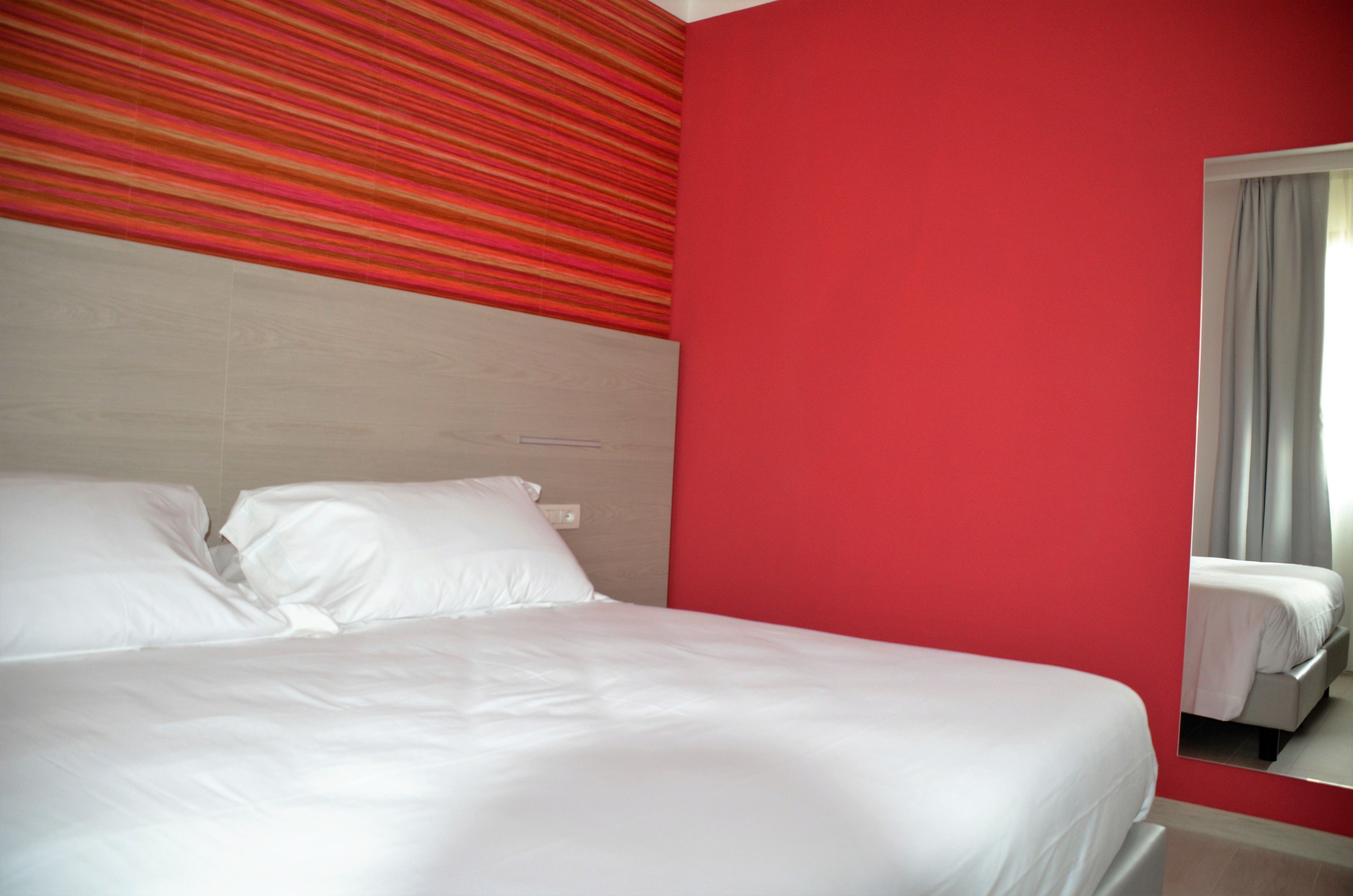 A hotel room with a bed, a red accent wall, and a window with curtains.