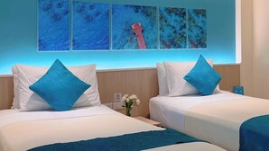 Habitación de hotel con dos camas, cada una con ropa blanca y almohadas azules. Hay una obra de arte colgada en la pared que representa un paisaje marino azul con un bote rojo.