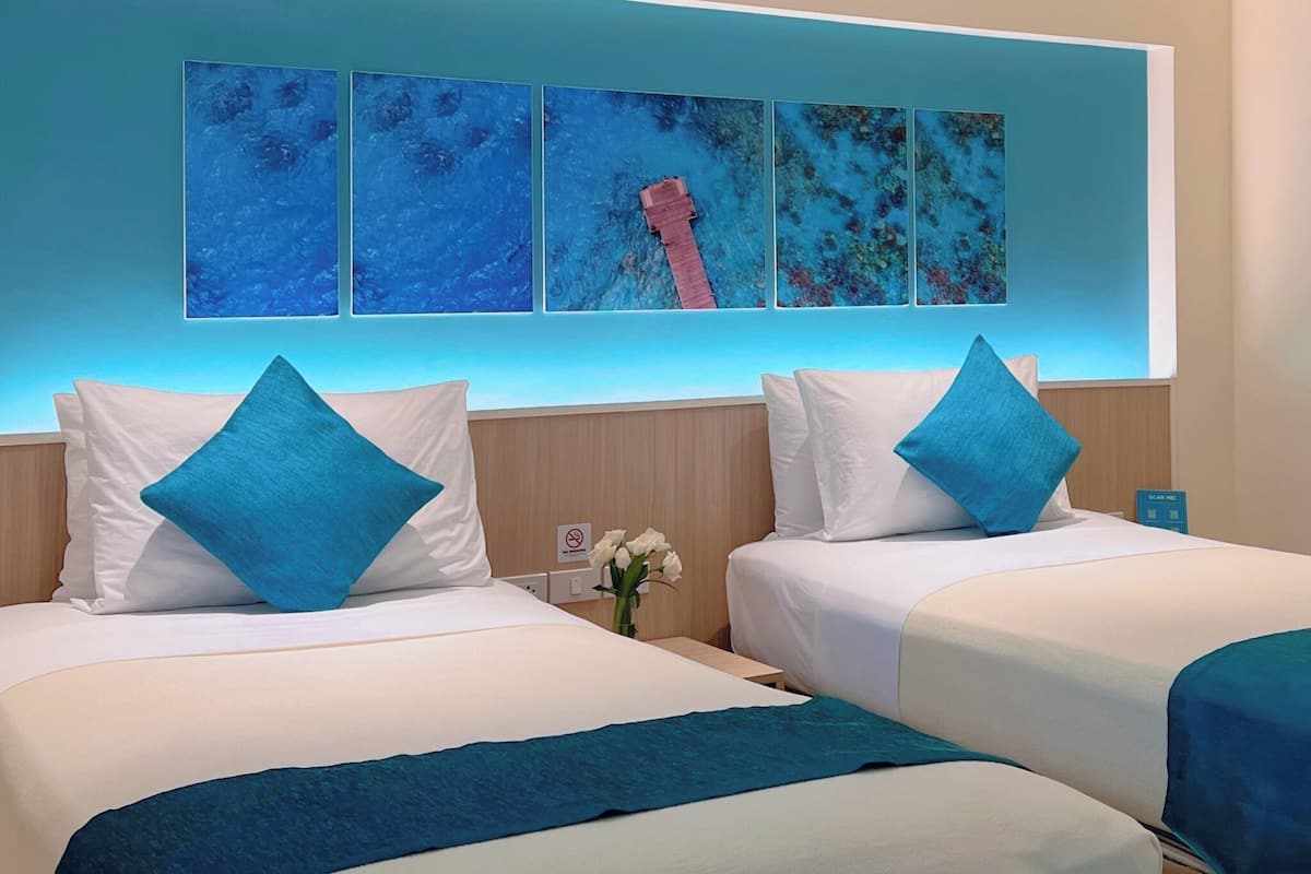 Ein Hotelzimmer mit zwei Betten, jeweils mit weißen Laken und blauen Kissen. An der Wand hängt ein Kunstwerk, das eine blaue Meereslandschaft mit einem roten Boot zeigt.