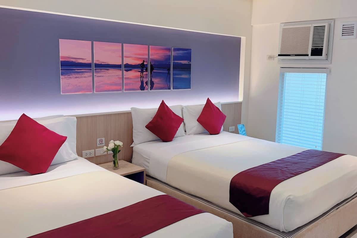 Ein Hotelzimmer mit zwei Betten, roten Kissen, einem Nachttisch mit Blumenvase und einer Wandklimaanlage.