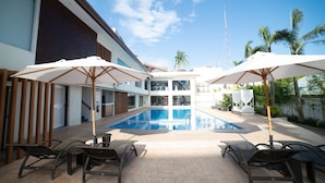 Zona de piscina con tumbonas y sombrillas, rodeada por un edificio moderno con grandes ventanales y vistas a palmeras.