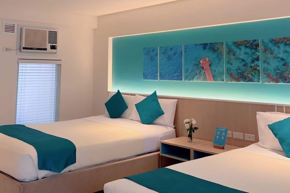 Ein Hotelzimmer mit zwei Betten, jeweils mit türkisfarbenem Bettzeug und Kissen, einem kleinen Nachttisch mit Blumenvase und einer wandmontierten Klimaanlage.