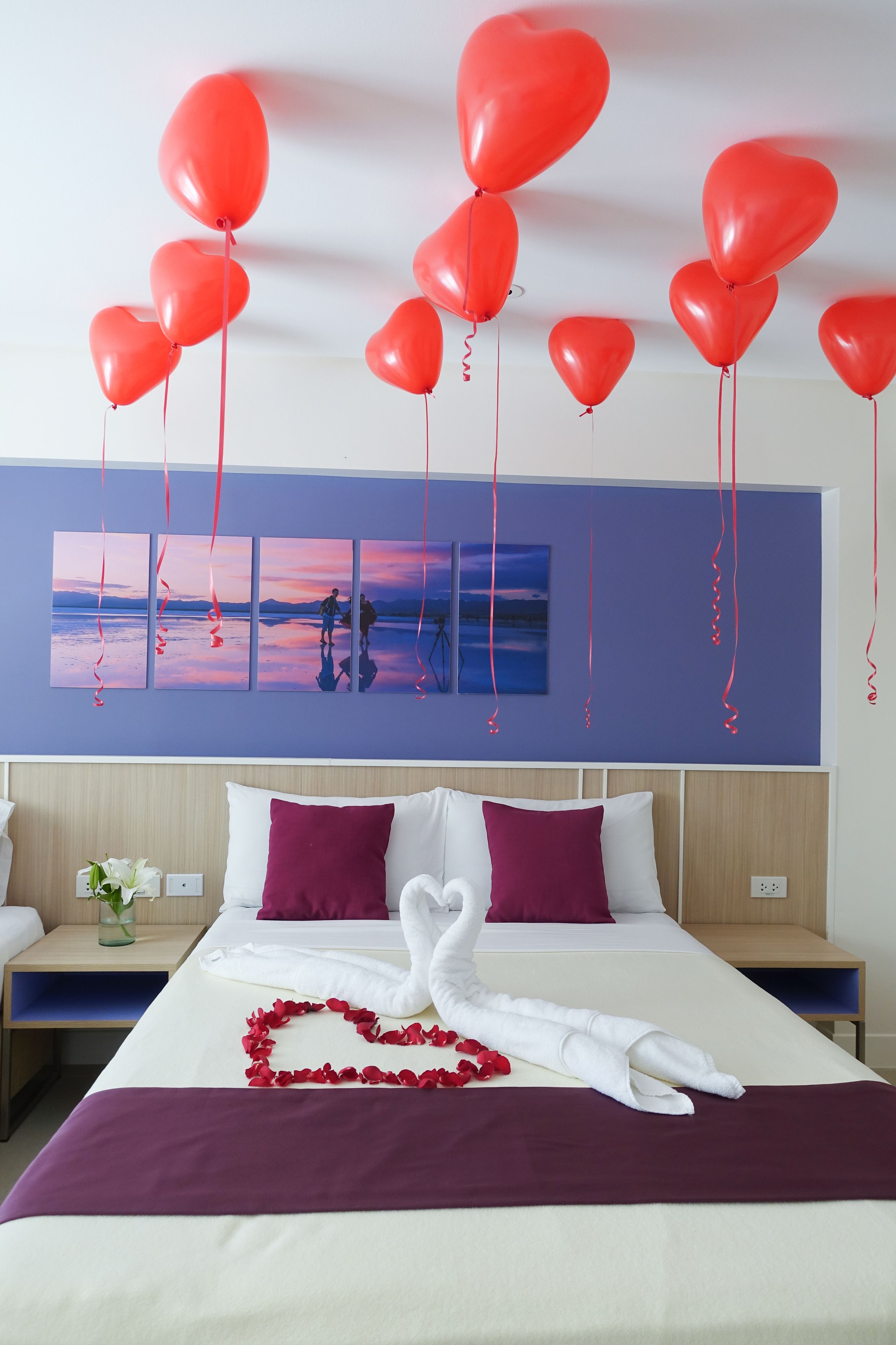 A hotel room with a bed, pillows, a swan-shaped towel, red heart-shaped balloons, and a sunset painting on the wall.