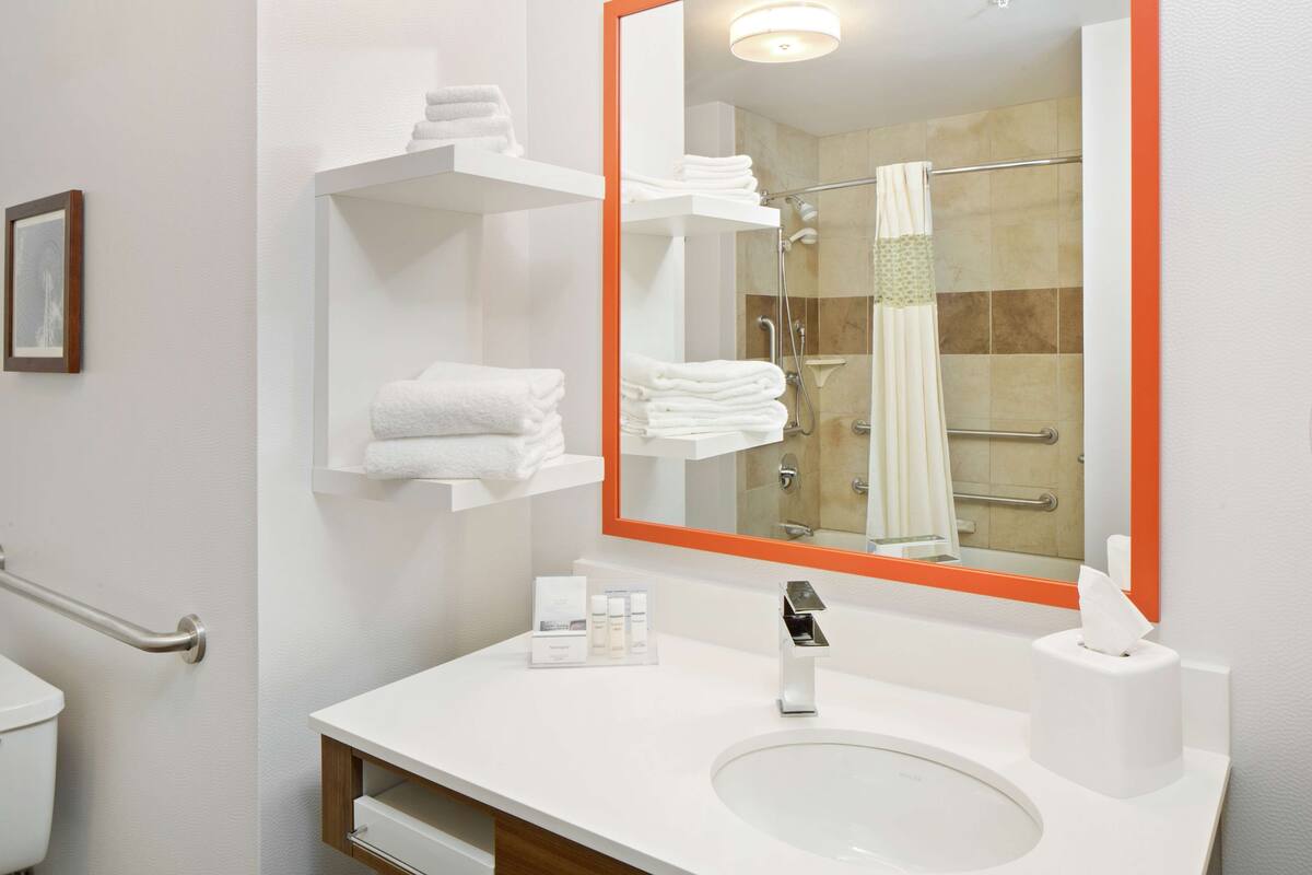 A modern bathroom with a white sink, a large mirror with an orange frame, and neatly stacked white towels on shelves.