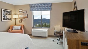 A hotel room with a bed, a desk with a flat-screen TV, a chair, a window with a view of mountains, and a wall-mounted air conditioner. - Hampton Inn & Suites Spokane Valley (Spokane Valley)