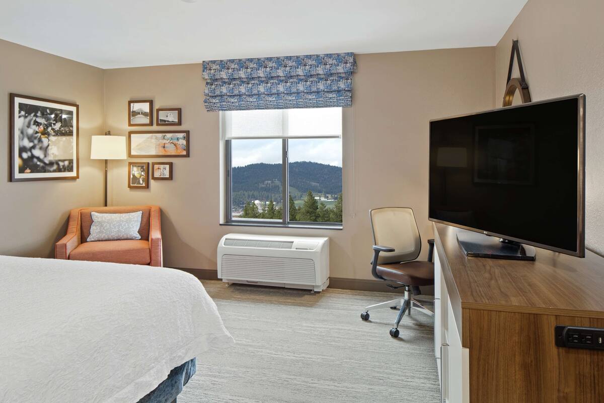A hotel room with a bed, a desk with a flat-screen TV, a chair, a window with a view of mountains, and a wall-mounted air conditioner.
