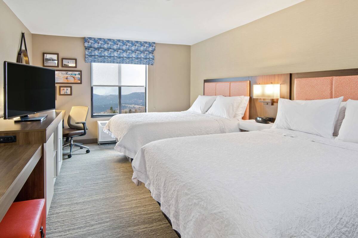 A hotel room with two beds, a desk with a television, a chair, and a window with a view of the mountains.