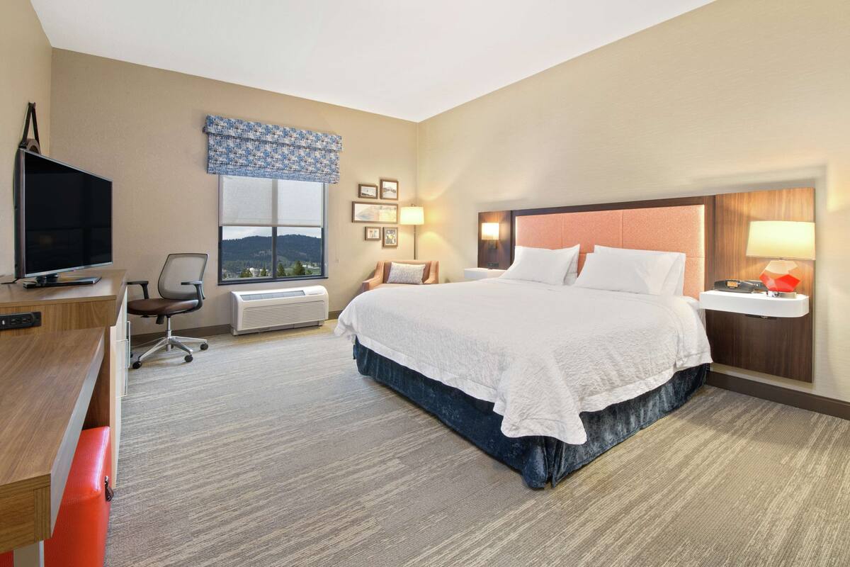 A hotel room with a large bed, a desk with a television, a chair, a window with a view, and a wall-mounted lamp.