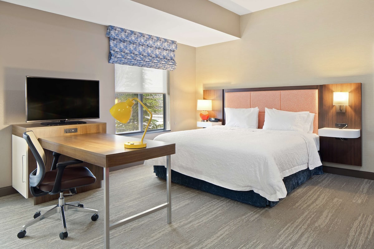 A hotel room with a bed, a desk with a TV, a lamp, and a window with a view.