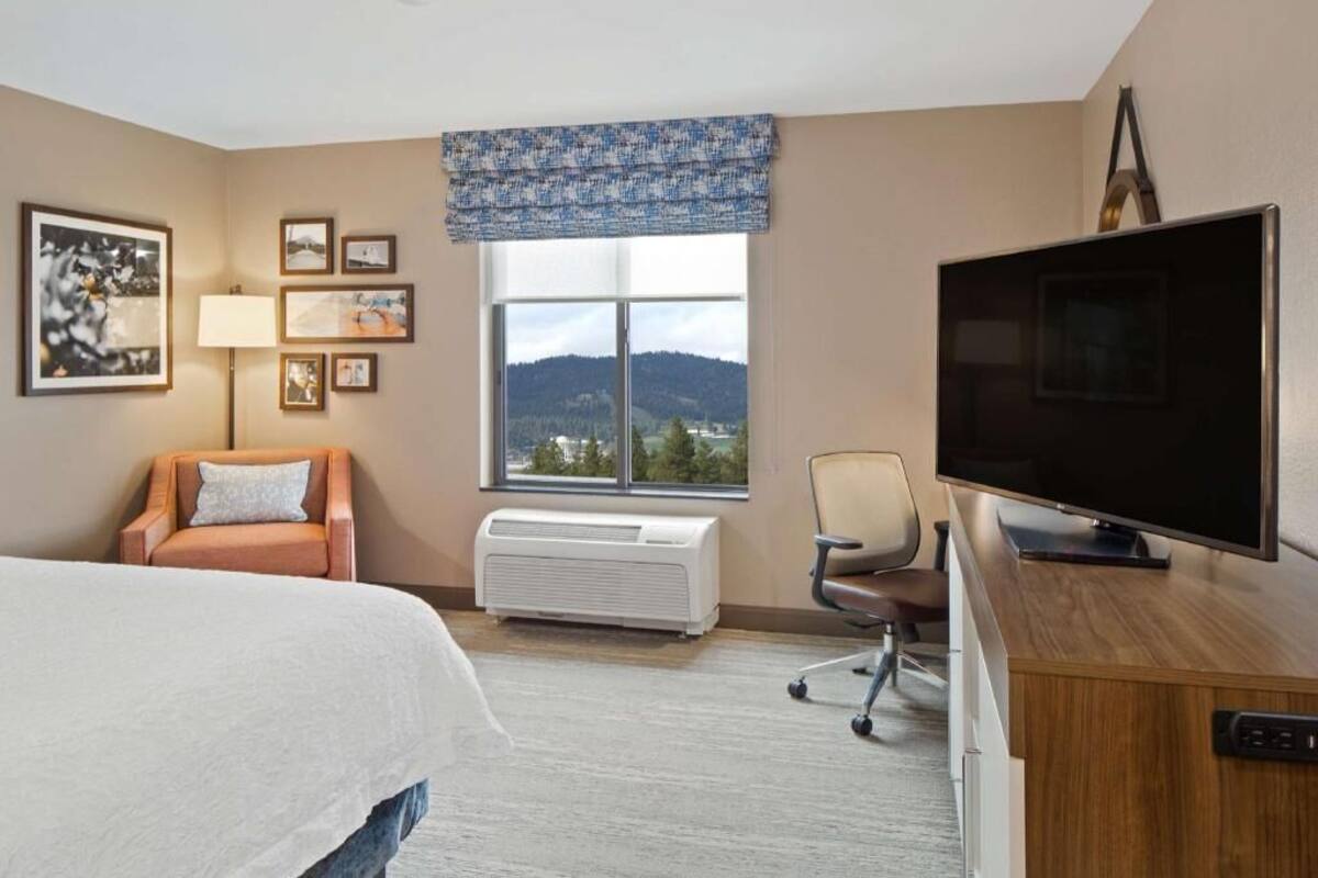 A hotel room with a bed, a desk with a flat-screen TV, a chair, a window with a view of mountains, and a wall-mounted air conditioner.