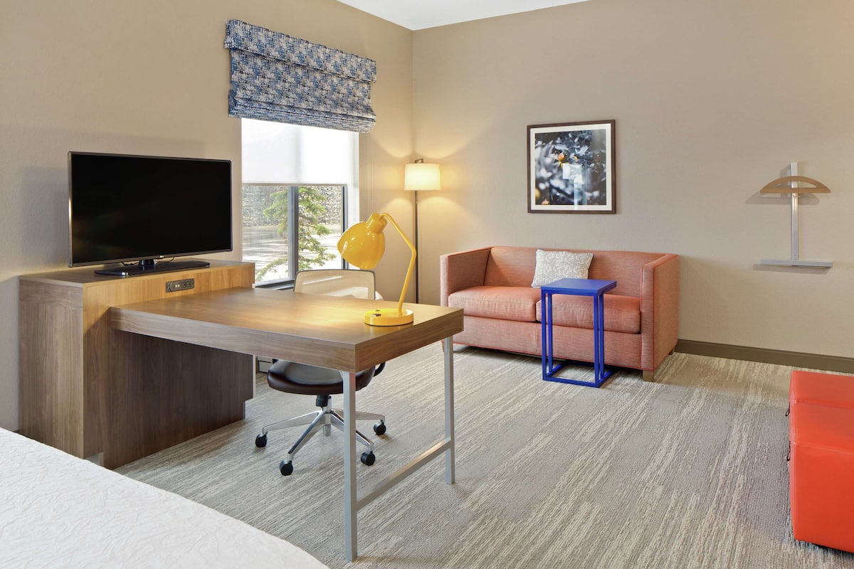 A hotel room with a wooden desk, a television, a sofa, a lamp, and a framed picture on the wall.