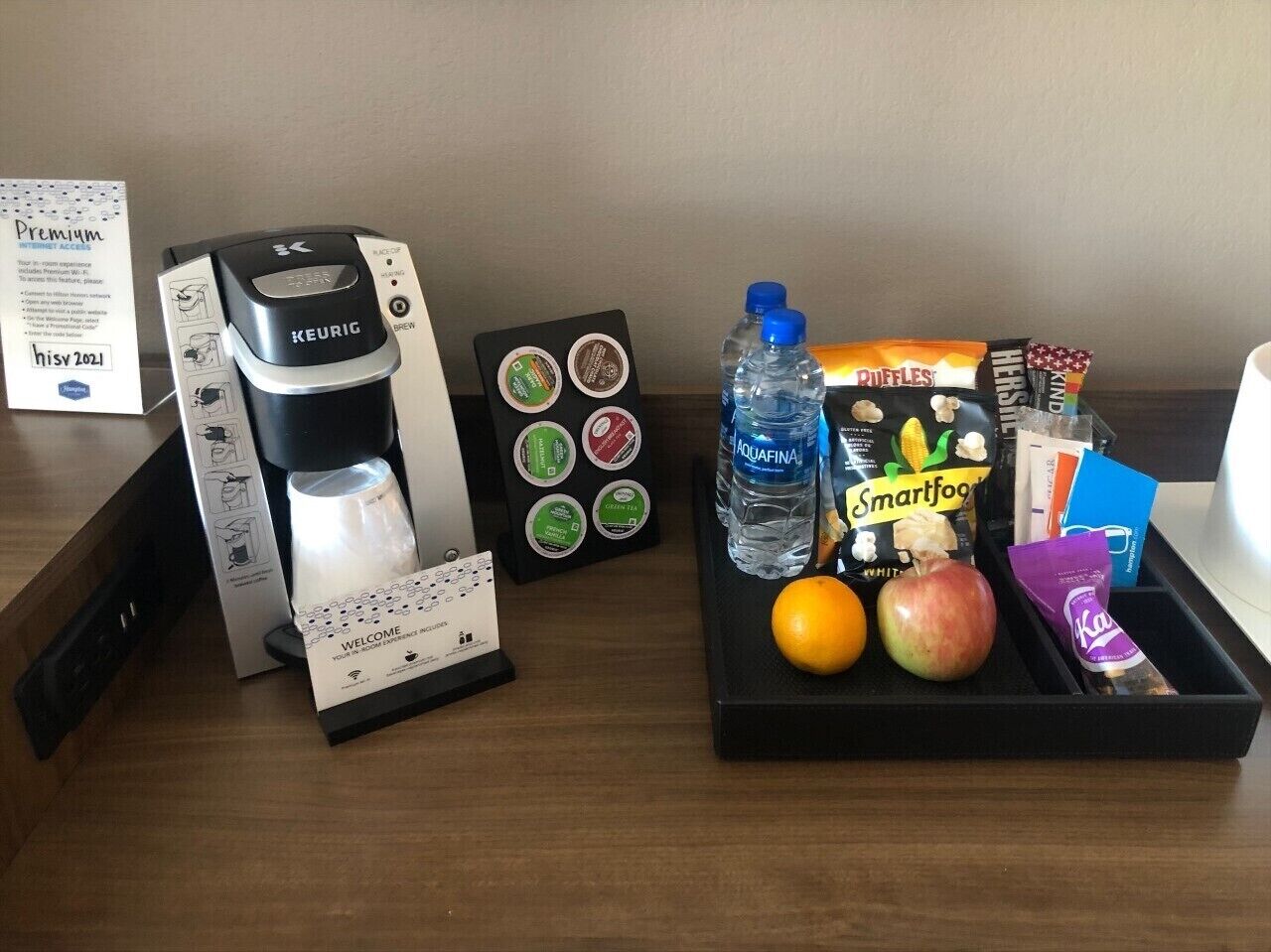 Una cafetera, una botella de agua, aperitivos y una cesta de fruta sobre una mesa de habitación de hotel.