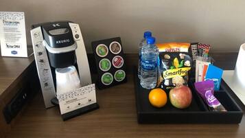 Una cafetera, una botella de agua, aperitivos y una cesta de fruta sobre una mesa de habitación de hotel.