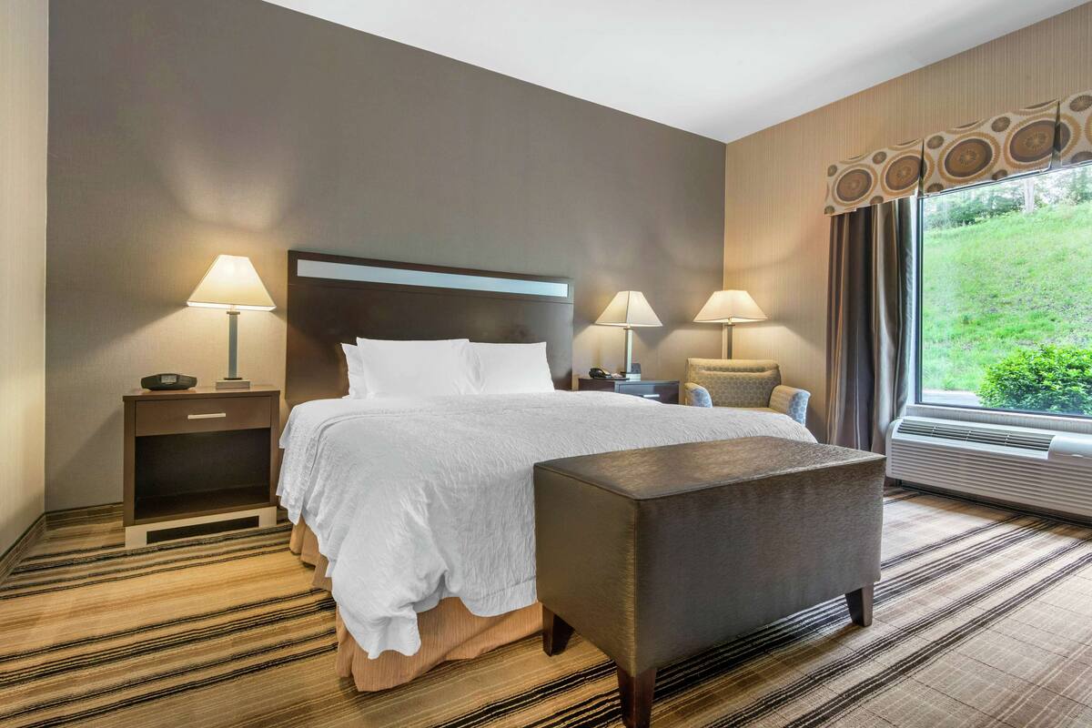 Habitación de hotel con una cama grande, dos mesitas de noche con lámparas, un banco, una ventana con cortinas y vistas a zonas verdes.