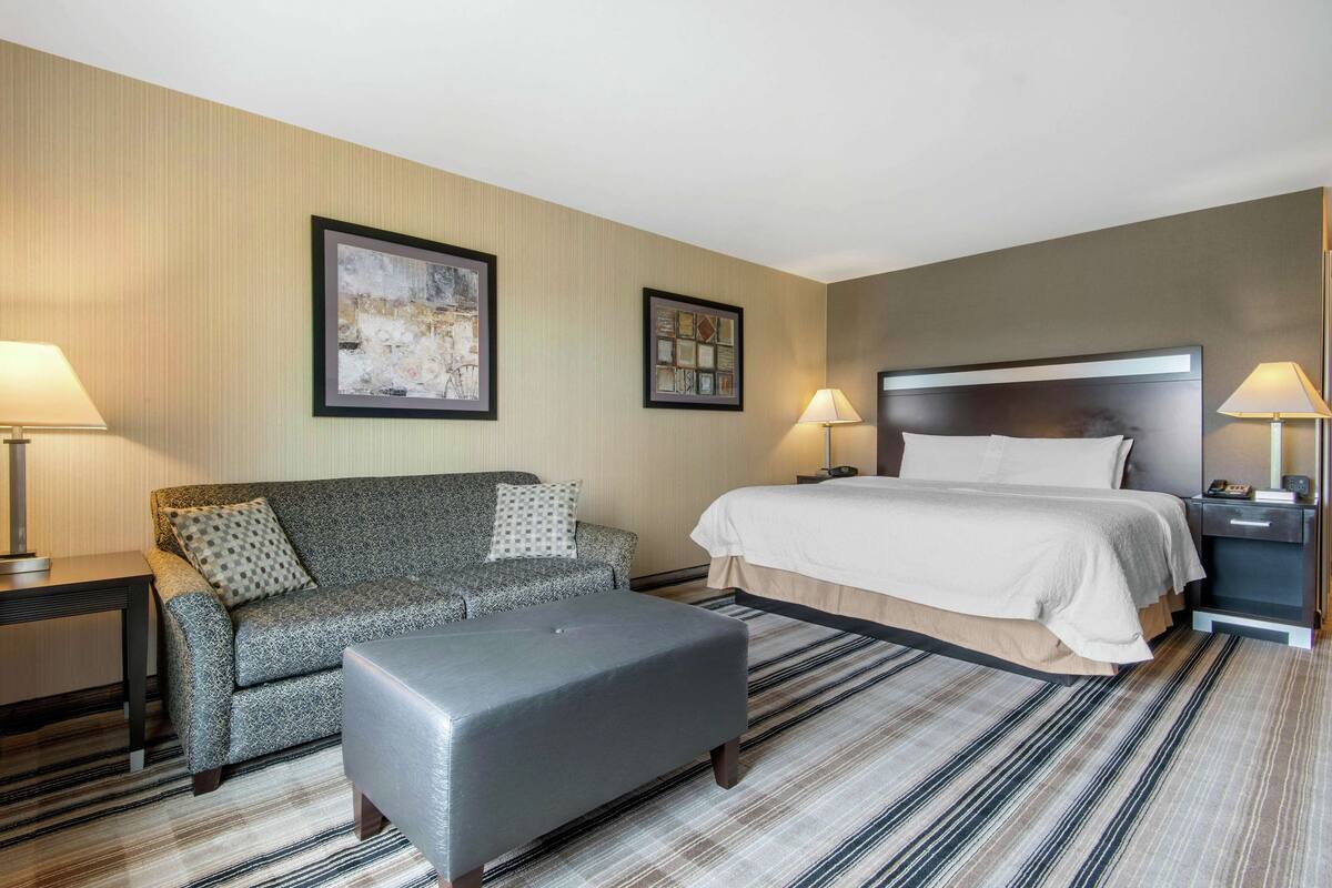 Una habitación de hotel con una cama, un sofá, un reposapiés y dos mesitas de noche, además de dos lámparas.