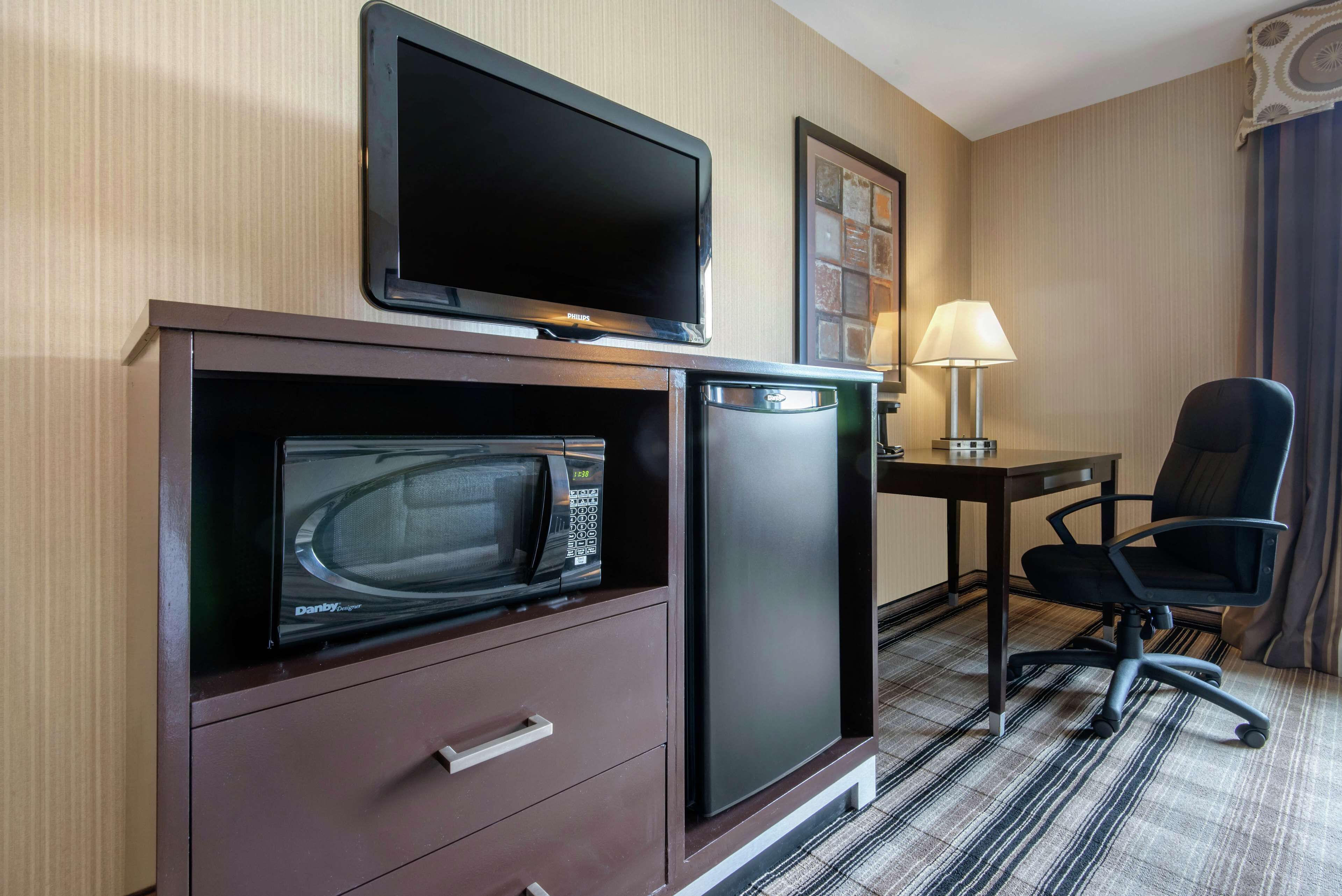 A compact hotel room with a flat-screen TV, a microwave, a mini-fridge, a desk with a lamp, and a black office chair.