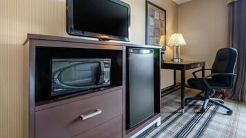 A compact hotel room with a flat-screen TV, a microwave, a mini-fridge, a desk with a lamp, and a black office chair.