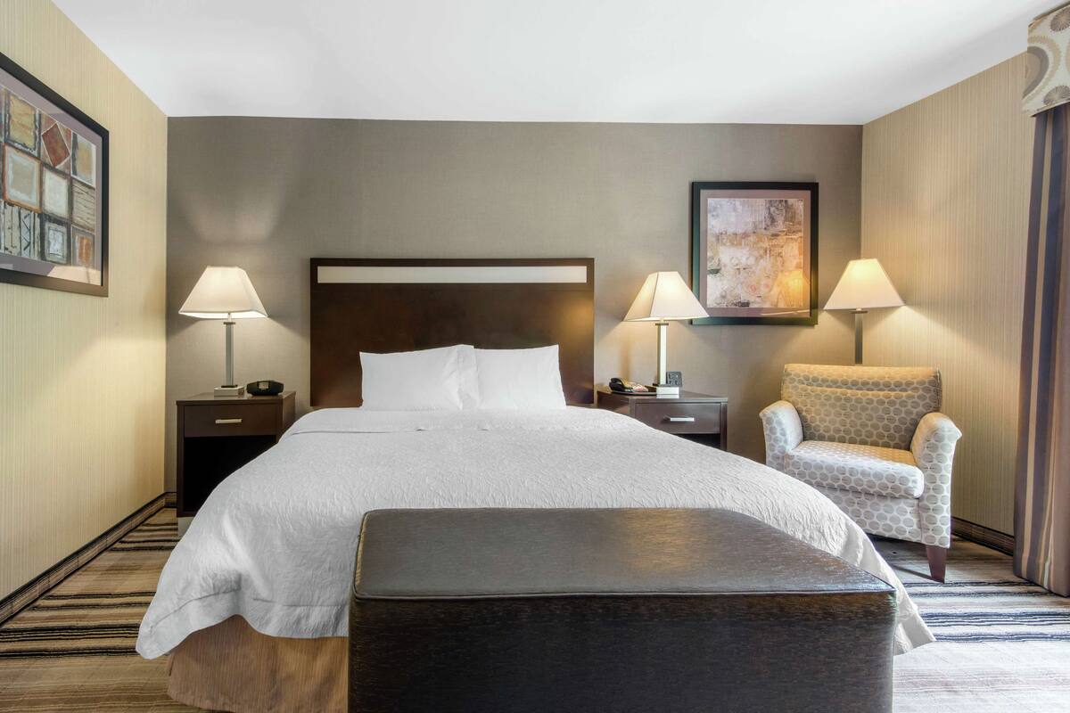 Una habitación de hotel con una cama grande, dos mesitas de noche, una silla y un banco.