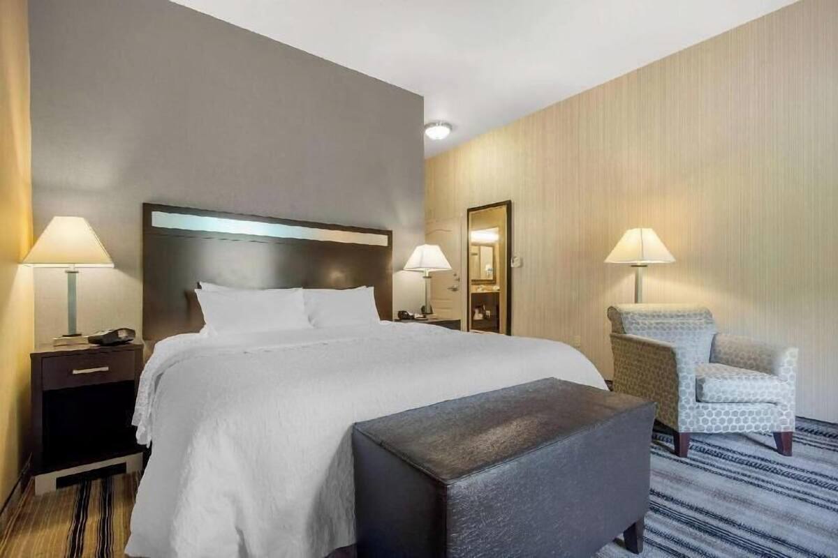 Una habitación de hotel con una cama, dos mesitas de noche con lámparas, una silla y un espejo.