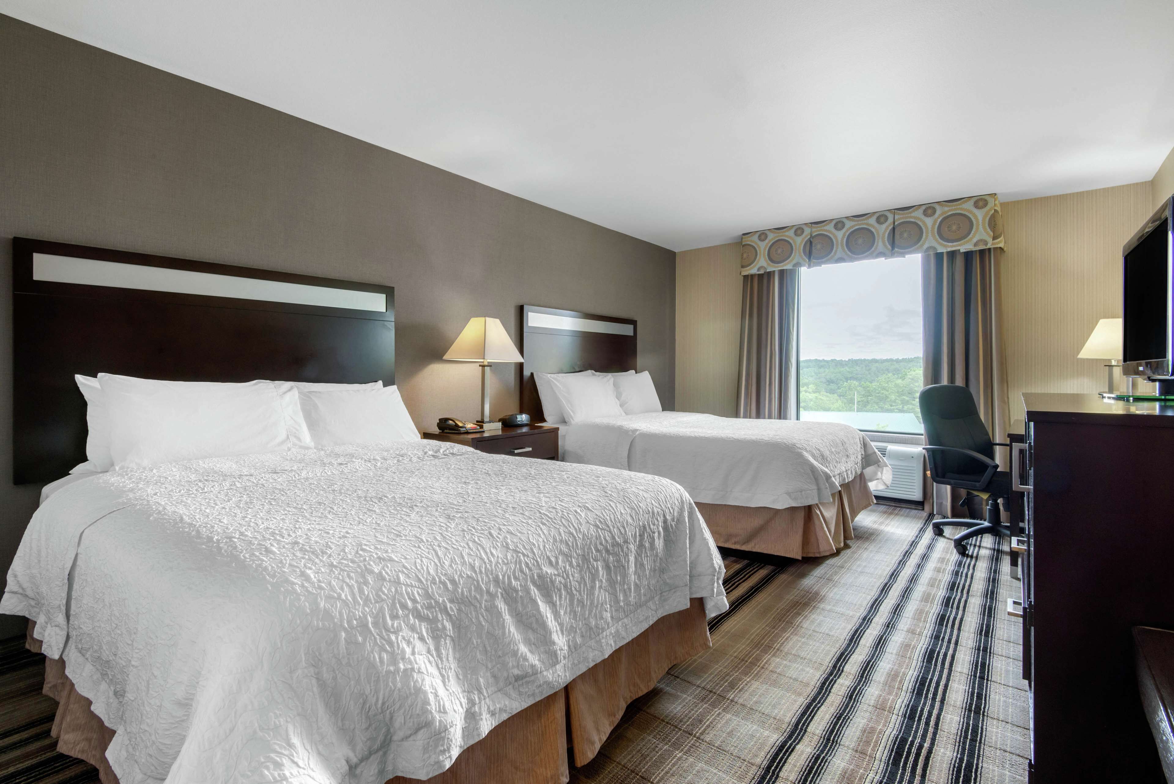 A hotel room with two beds, a desk, a chair, and a large window with a view.