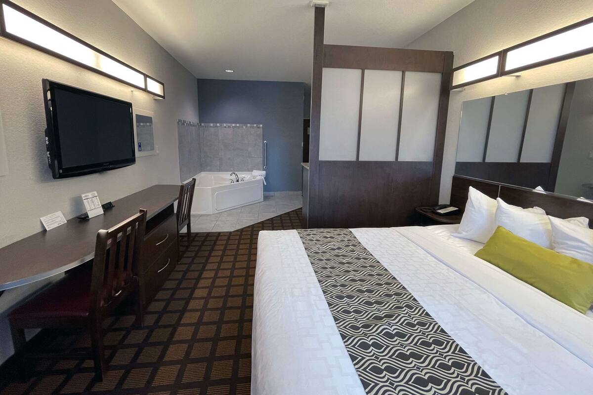 A hotel room with a bed, a desk, a chair, a TV, a bathroom with a bathtub, and a large mirror.