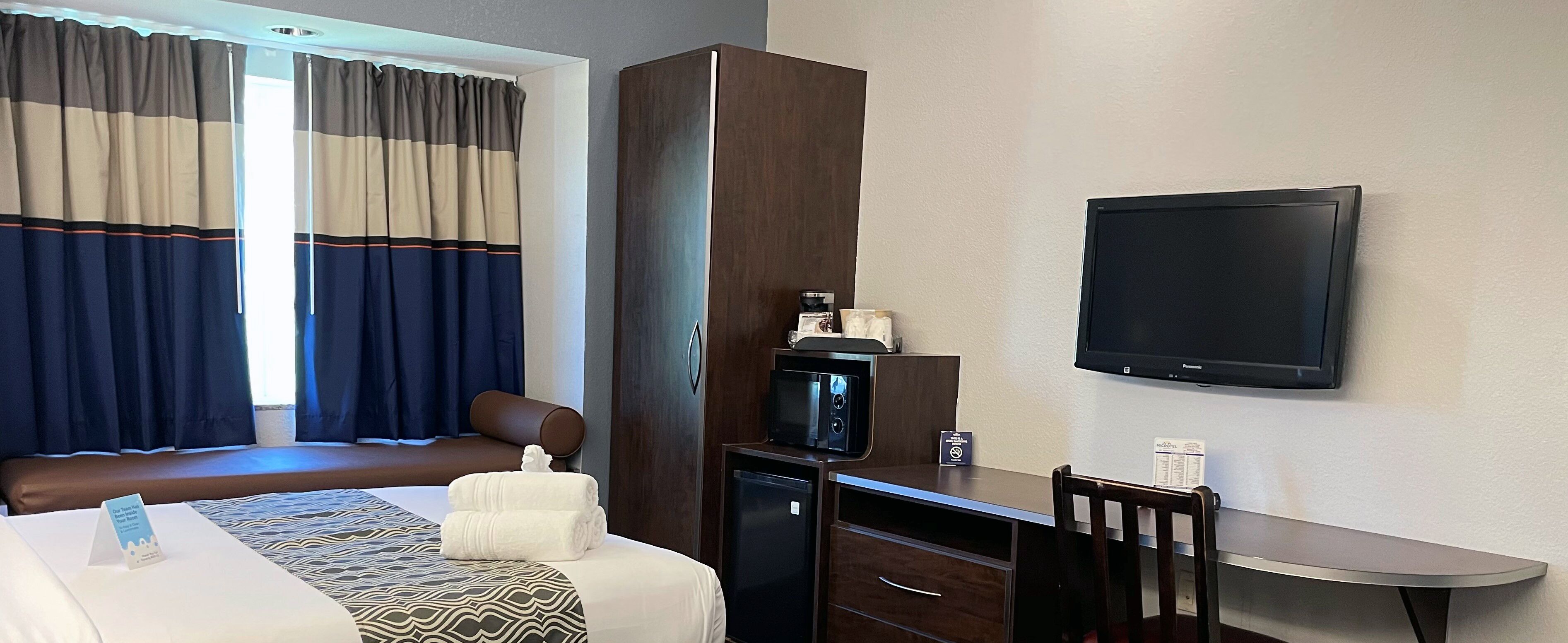 Ett hotellrum med en säng, ett skrivbord med en stol, en tv, en mikrovågsugn och ett litet kylskåp.