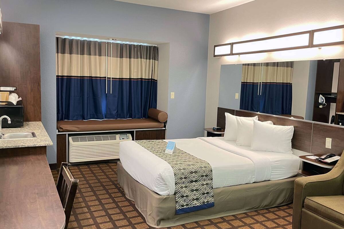 A hotel room with a bed, a desk with a sink, a chair, a window with curtains, and a mirror.