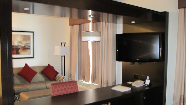 Habitación de hotel con cama, escritorio con teléfono, televisor, sofá con cojines rojos, lámpara y un cuadro en la pared.