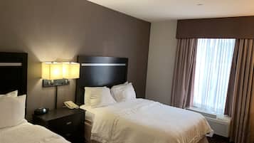 A hotel room with two beds, a bedside table with a lamp, a phone, and a window with curtains.