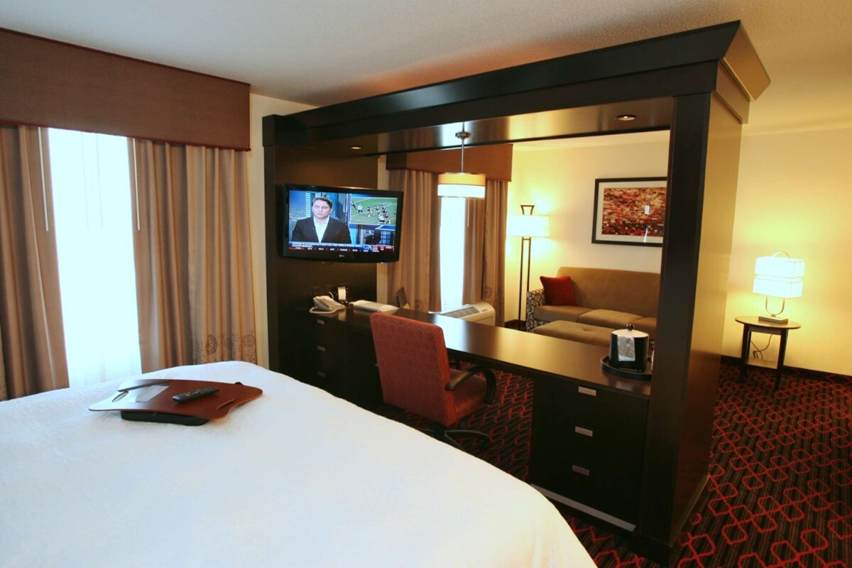 A hotel room with a large bed, a desk with a TV, a desk lamp, a chair, a sofa, and a carpeted floor.