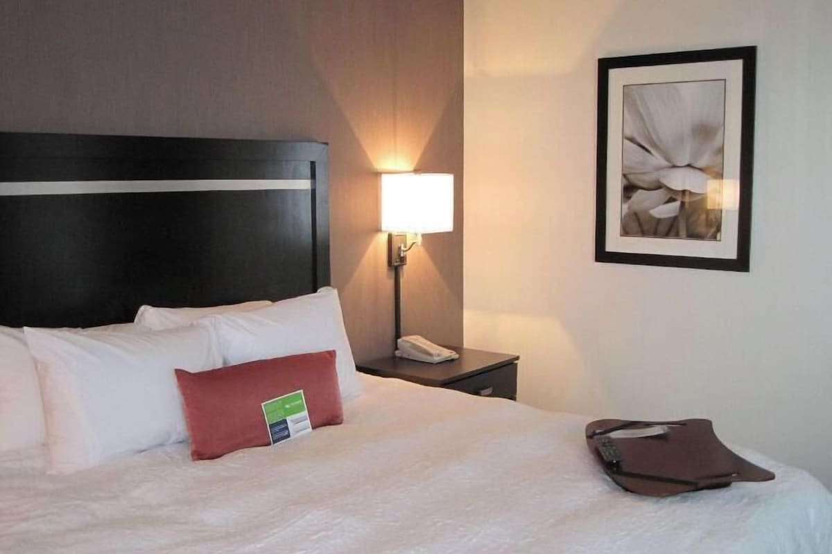 A hotel room with a bed, a nightstand with a lamp, a framed picture on the wall, and a brown bag on the bed.