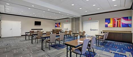 A hotel conference room with multiple tables and chairs, a carpeted floor, and a ceiling with recessed lighting.