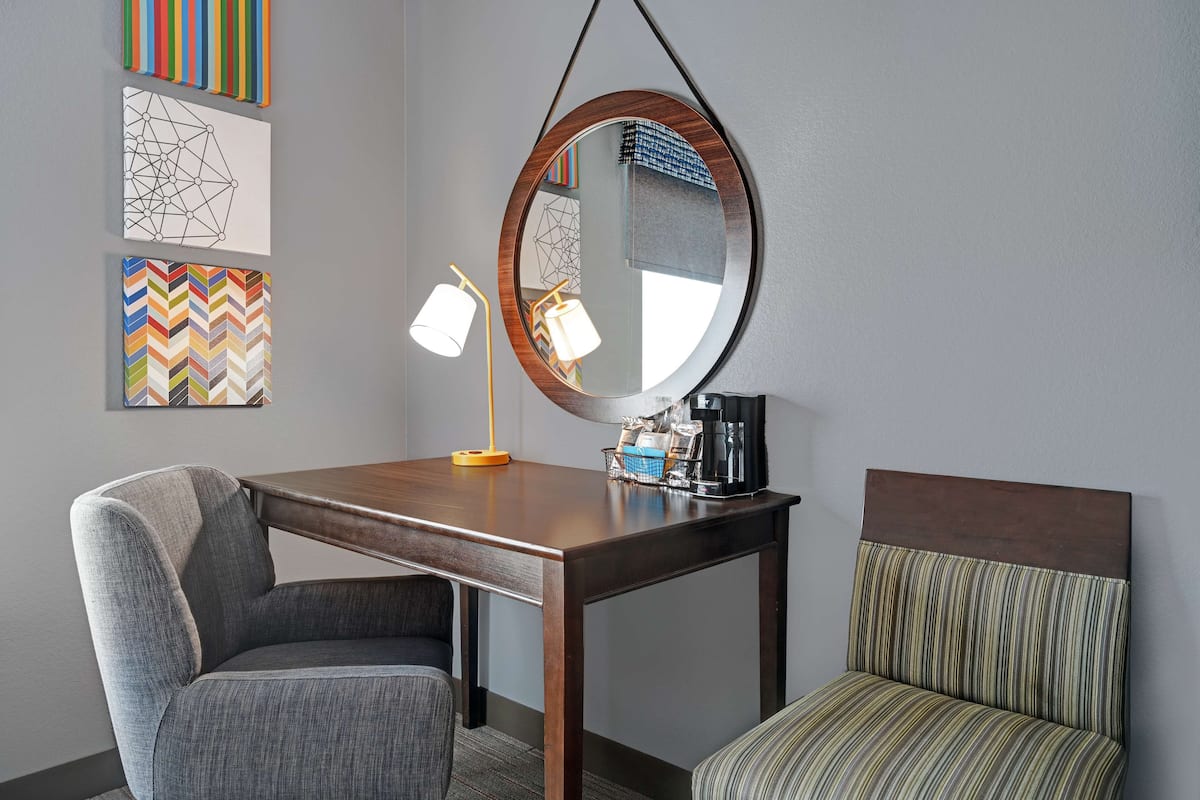 A hotel room with a wooden table, a chair, a sofa, a round mirror, and artwork on the wall.