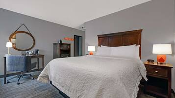 A hotel room with a large bed, two bedside lamps, a desk with a chair, a microwave, and a small clock on the nightstand.