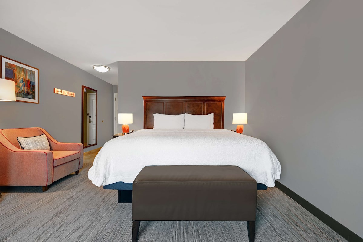 A hotel room with a large bed, a brown bench, a red armchair, and two bedside lamps.