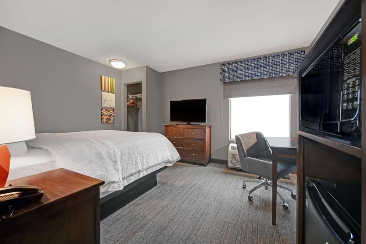 A hotel room with a bed, a desk with a chair, a television, a dresser, and a large window.