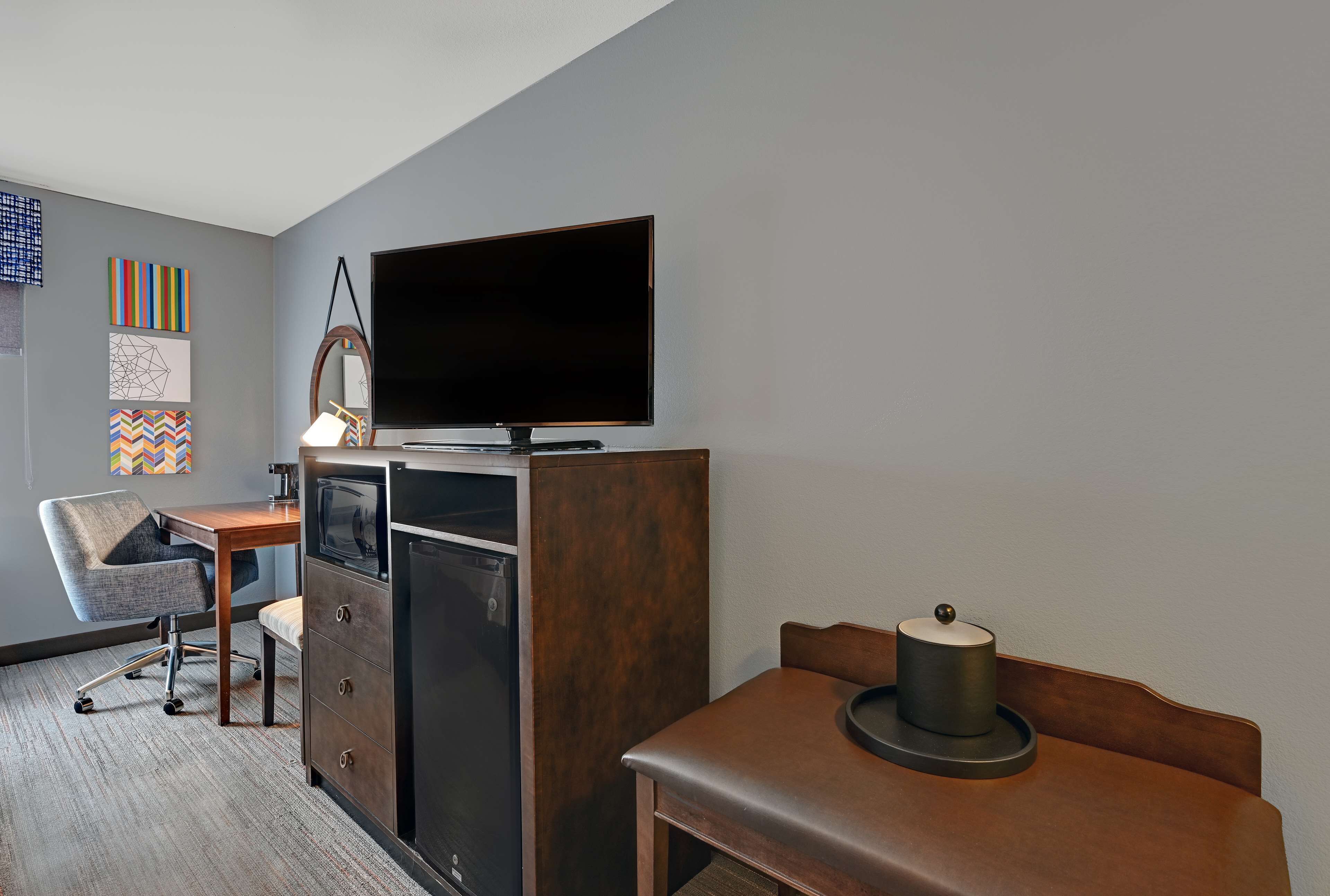 Ein Hotelzimmer mit Flachbildfernseher an einem Holzkasten, einem Schreibtisch mit Stuhl, einem kleinen Kühlschrank und einem Nachttisch mit Lampe.