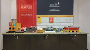 A kitchen counter with waffle-making equipment, waffle boxes, and a sign promoting waffles.