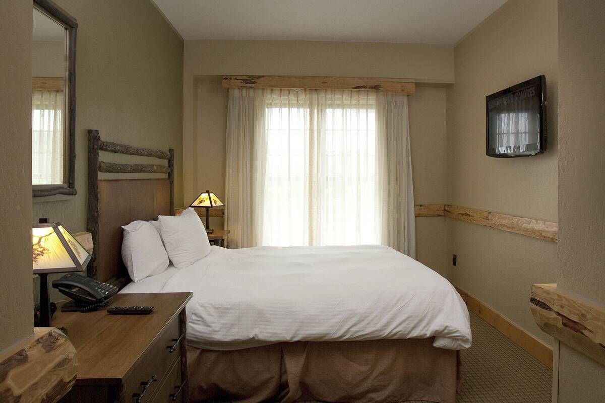 A hotel room with a large bed, a desk with a lamp, a telephone, and a television mounted on the wall.