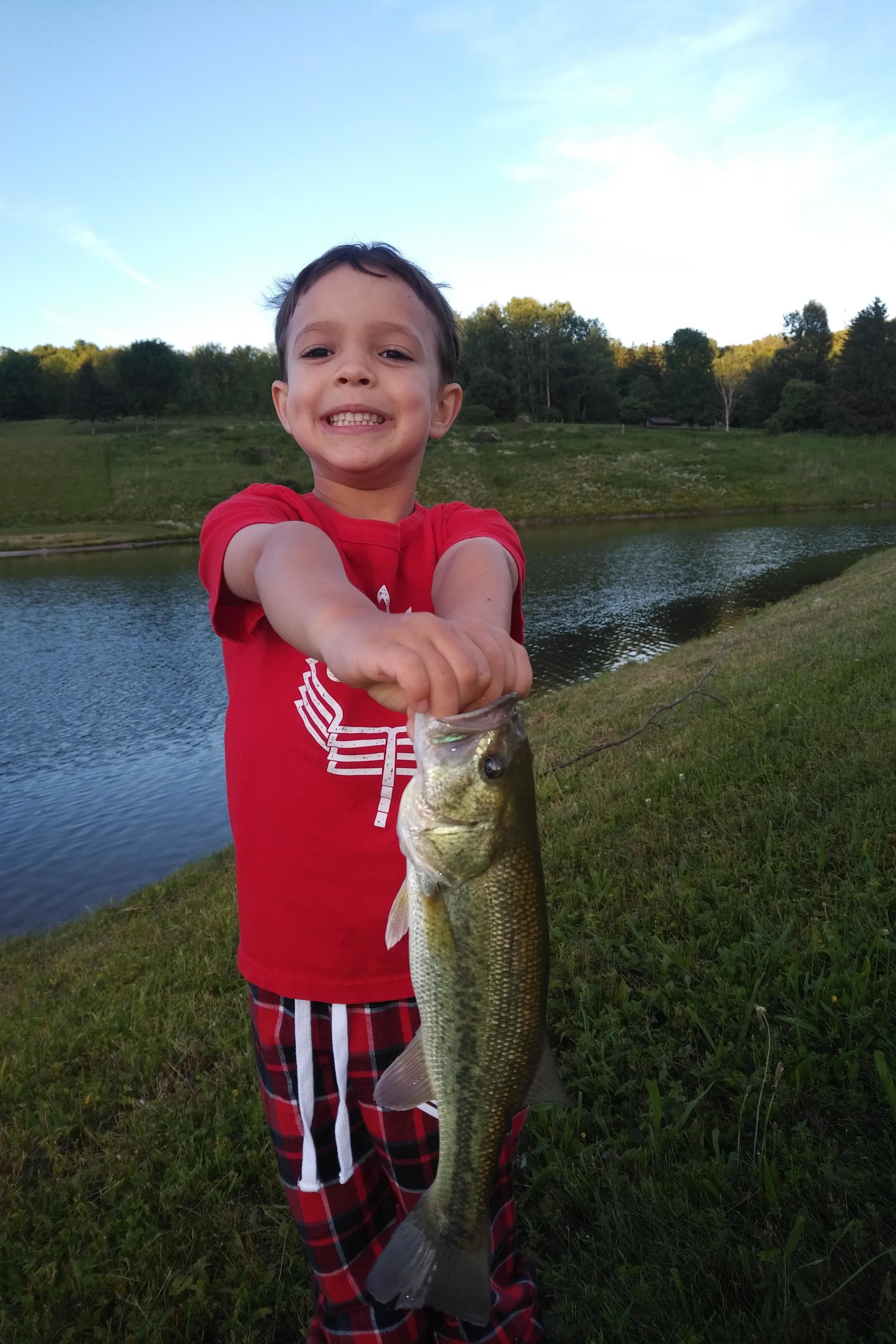 A child holding a fish by a body of water with trees and a building in the background.