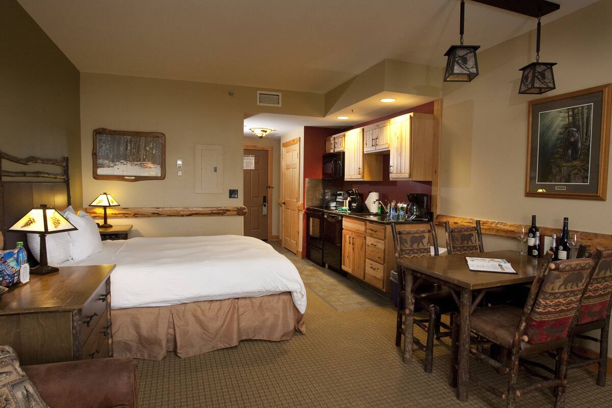 A hotel room with a bed, a desk with a lamp, a dining table with chairs, a kitchen area, and a painting on the wall.