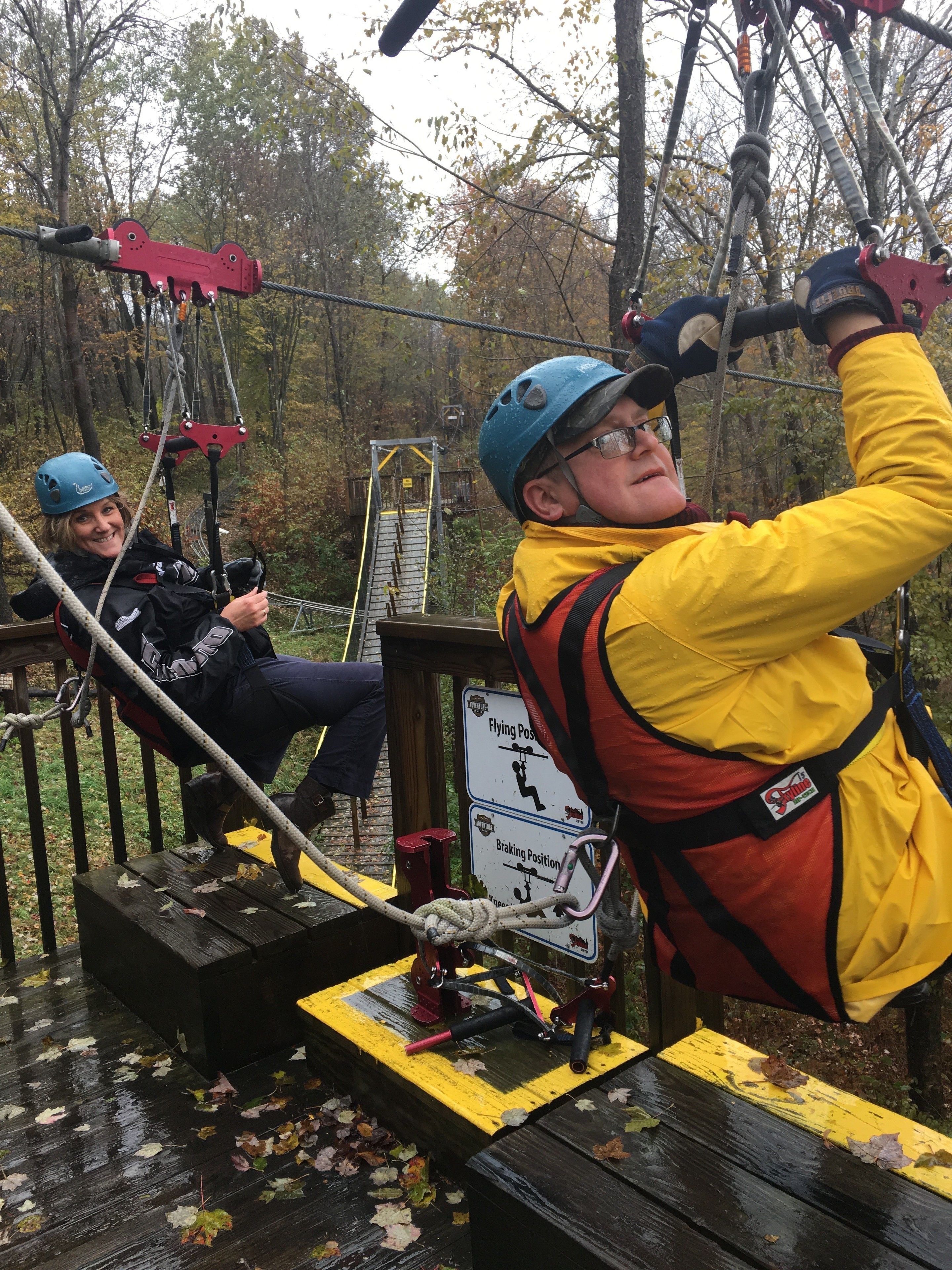 A zip-lining activity with participants wearing helmets and harnesses, set in a wooded area with a wooden platform and safety instructions visible.