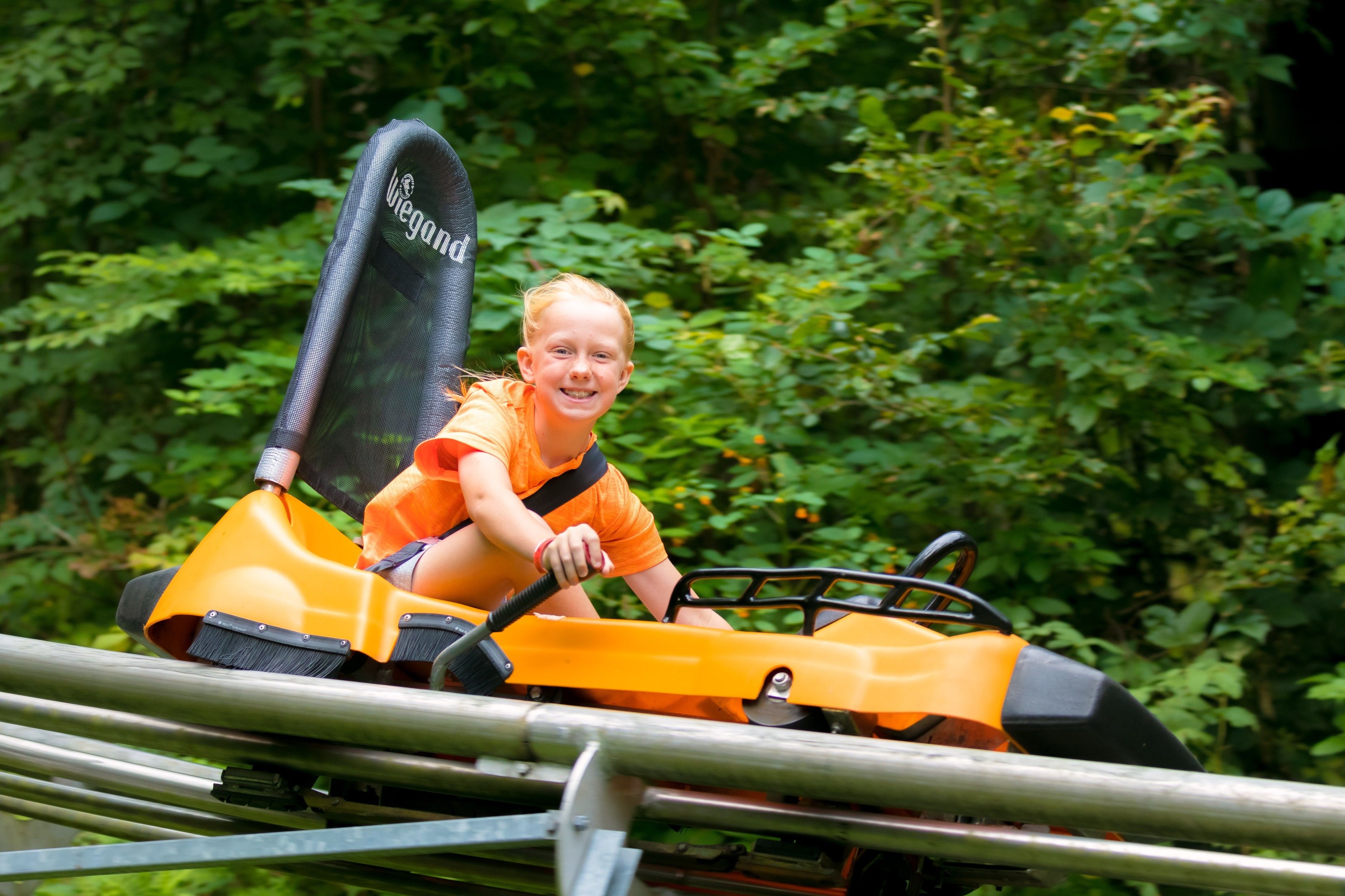 A child in an orange ride-on vehicle with a safety harness, surrounded by greenery.
