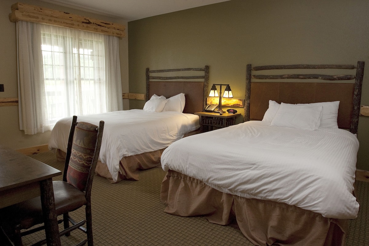 A hotel room with two single beds, a wooden nightstand with a lamp, a wooden desk, and a chair.